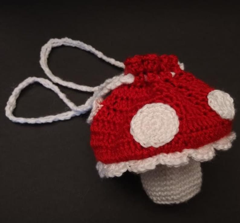 May include: A crocheted red and white mushroom-shaped bag with a white drawstring and white polka dots. The bag has a white crocheted stem.