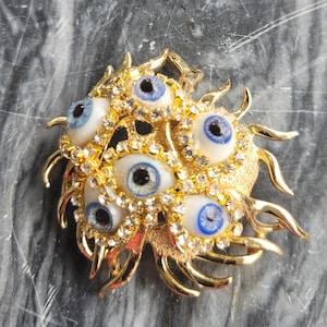 May include: A gold-toned brooch featuring multiple blue-eyed, white-irised eyes, surrounded by clear crystals and gold accents. The brooch has a sunburst design with curved, pointed rays extending outwards. A unique, eye-catching accessory.