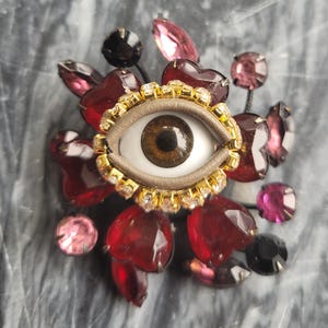 May include: A decorative brooch featuring a central eye design with a brown iris, surrounded by a gold-colored frame with clear rhinestones. The brooch is adorned with red heart-shaped and pink gemstone accents.