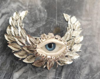 May include: Silver-toned brooch featuring a detailed eye with a blue iris, set within a white fabric. The eye is surrounded by a wreath of silver leaves and sparkling crystals. A unique and decorative accessory.