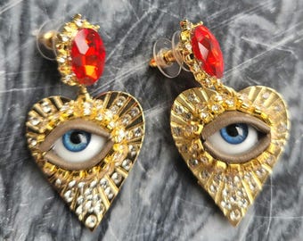 May include: A pair of gold-toned heart-shaped earrings. Each earring features a blue eye, surrounded by a gold frame with clear crystals. A red gemstone tops each earring. The earrings have a post-back closure.