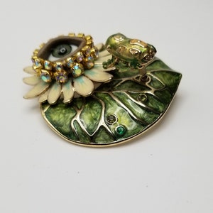 Pode incluir: Um broche dourado com uma flor branca com um olho verde no centro, rodeado por strass. Um sapo verde est&aacute; sentado em uma folha verde com detalhes dourados.