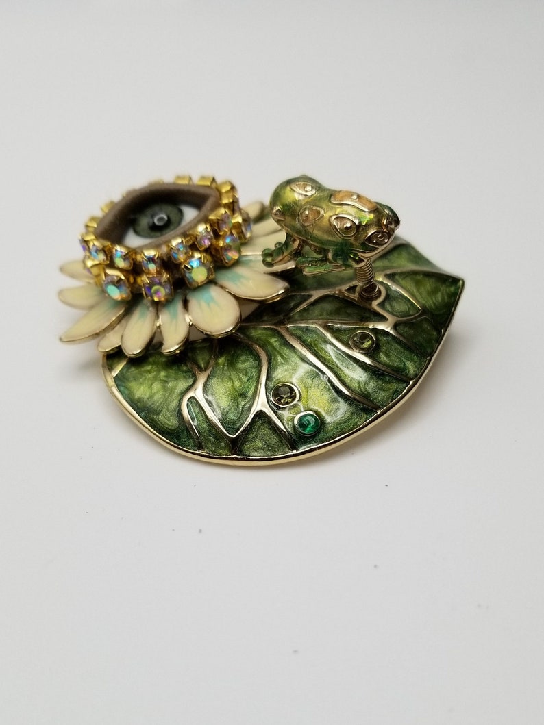 Pode incluir: Um broche dourado com uma flor branca com um olho verde no centro, rodeado por strass. Um sapo verde est&aacute; sentado em uma folha verde com detalhes dourados.