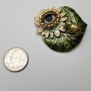 Pode incluir: Um broche dourado com um design de esmalte verde e branco. O broche apresenta um olho grande com uma &iacute;ris marrom e uma pupila verde, rodeado por uma flor branca com detalhes dourados. Um pequeno sapo verde est&aacute; empoleirado na folha.