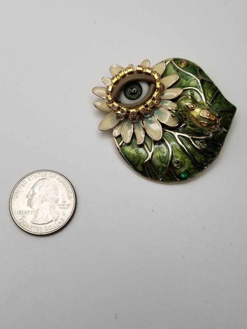 Pode incluir: Um broche dourado com um design de esmalte verde e branco. O broche apresenta um olho grande com uma &iacute;ris marrom e uma pupila verde, rodeado por uma flor branca com detalhes dourados. Um pequeno sapo verde est&aacute; empoleirado na folha.