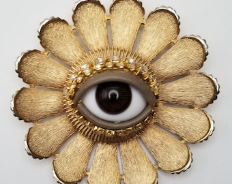 May include: A gold-toned flower brooch with a large, white eye in the center. The eye has a black pupil and is surrounded by a ring of rhinestones.