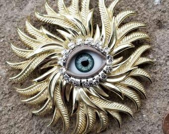 May include: A gold-toned brooch with a large, faux eye in the center. The eye is surrounded by a ring of clear rhinestones. The brooch is shaped like a sunburst with many points.