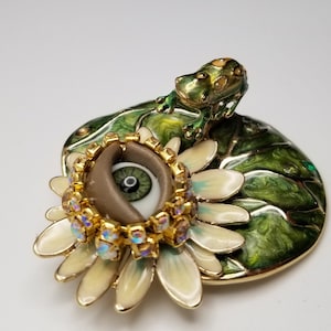 Pode incluir: Uma caixa de joias em tom dourado com uma tampa de esmalte verde que representa um l&iacute;rio d'&aacute;gua com um sapo no topo. A caixa abre para revelar uma flor branca com um olho castanho no centro, rodeado por strass dourados e iridescentes.