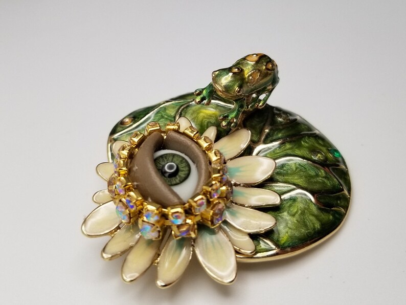 Pode incluir: Uma caixa de joias em tom dourado com uma tampa de esmalte verde que representa um l&iacute;rio d'&aacute;gua com um sapo no topo. A caixa abre para revelar uma flor branca com um olho castanho no centro, rodeado por strass dourados e iridescentes.