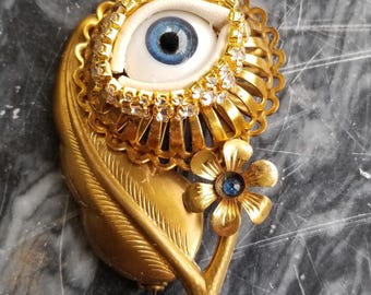 May include: A gold-toned brooch with a blue eye and a flower. The eye is surrounded by rhinestones and the flower has a blue center.