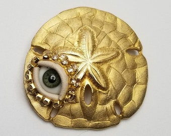 May include: A gold-coloured sand dollar brooch with a textured surface. The brooch features a decorative eye with a green iris, surrounded by small crystals and gold-coloured metal detailing. The brooch has a starfish design in the centre.