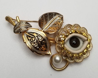 May include: A gold-tone metal brooch featuring a central eye motif with a white iris and dark pupil, surrounded by a scalloped frame. A single white pearl hangs below. Two engraved leaves with dark accents are attached to the stem of the brooch.