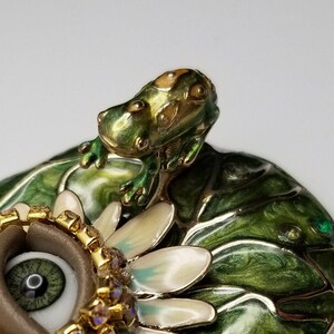 Pode incluir: Uma figura de sapo verde empoleirada em um l&iacute;rio d'&aacute;gua com uma flor branca e um olho marrom com detalhes dourados e de cristal.