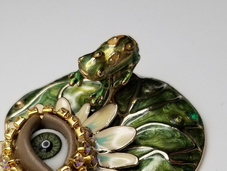 Pode incluir: Uma figura de sapo verde empoleirada em um l&iacute;rio d'&aacute;gua com uma flor branca e um olho marrom com detalhes dourados e de cristal.