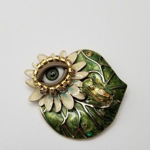 Pode incluir: Um broche de esmalte verde e dourado com uma flor branca com um olho grande, marrom e verde no centro. Um pequeno sapo verde est&aacute; sentado na flor.