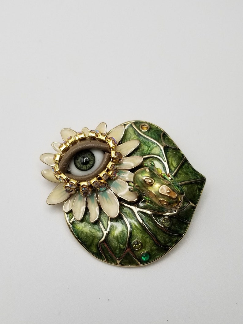 Pode incluir: Um broche de esmalte verde e dourado com uma flor branca com um olho grande, marrom e verde no centro. Um pequeno sapo verde est&aacute; sentado na flor.