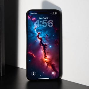 May include: A black smartphone displays a vibrant nebula wallpaper with shades of blue, red, and orange. The screen shows the time as 4:56 on Sunday, February 15. The phone is on a dark surface with a white wall in the background.