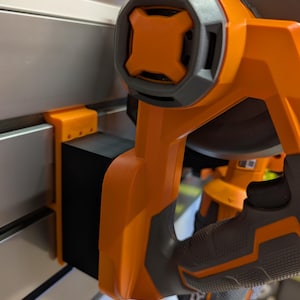 May include: Close-up of an orange and gray power tool. The tool has a black rectangular component attached to a metal surface. The handle is orange with a gray grip.
