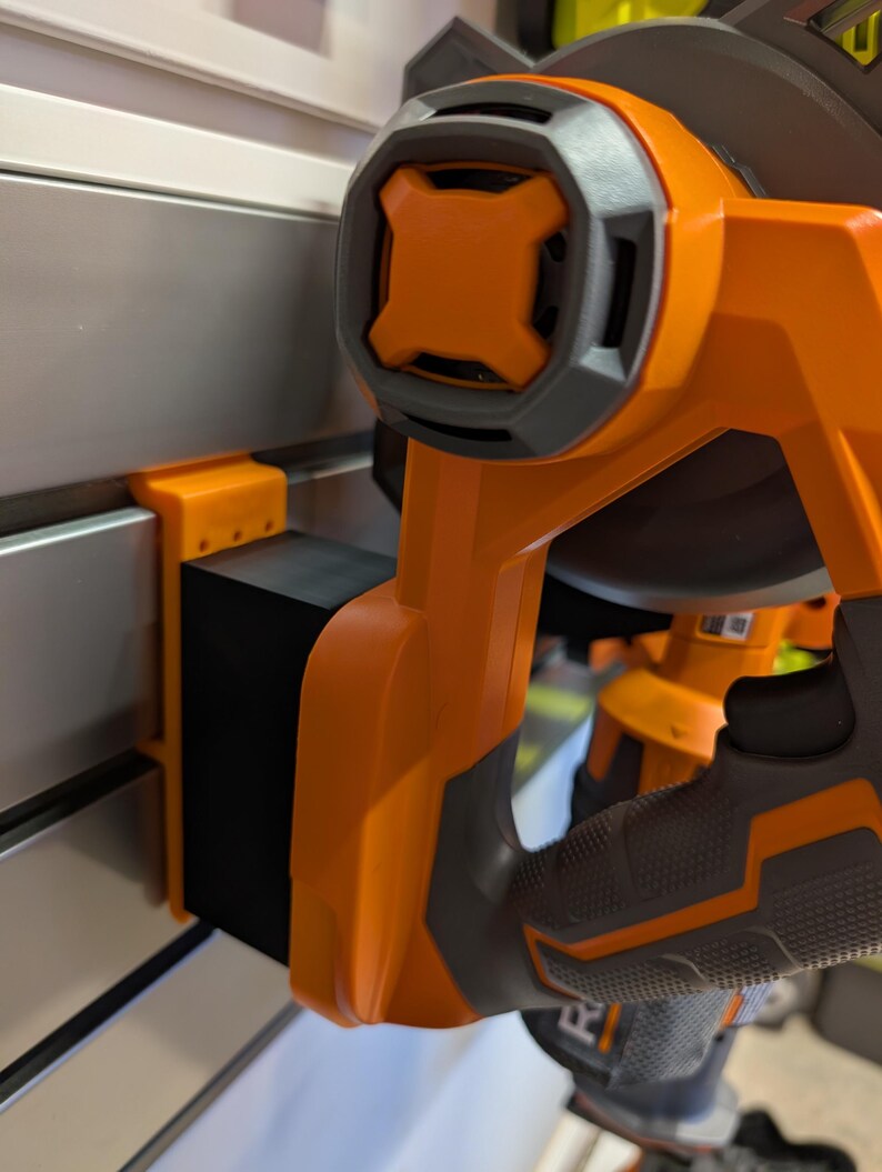 May include: Close-up of an orange and gray power tool. The tool has a black rectangular component attached to a metal surface. The handle is orange with a gray grip.