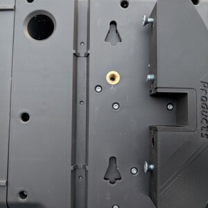 May include: Close-up of a black mounting bracket with the text "Peg d Products" printed on the side. The bracket is attached to a gray device with screws. The device has several holes and mounting points.