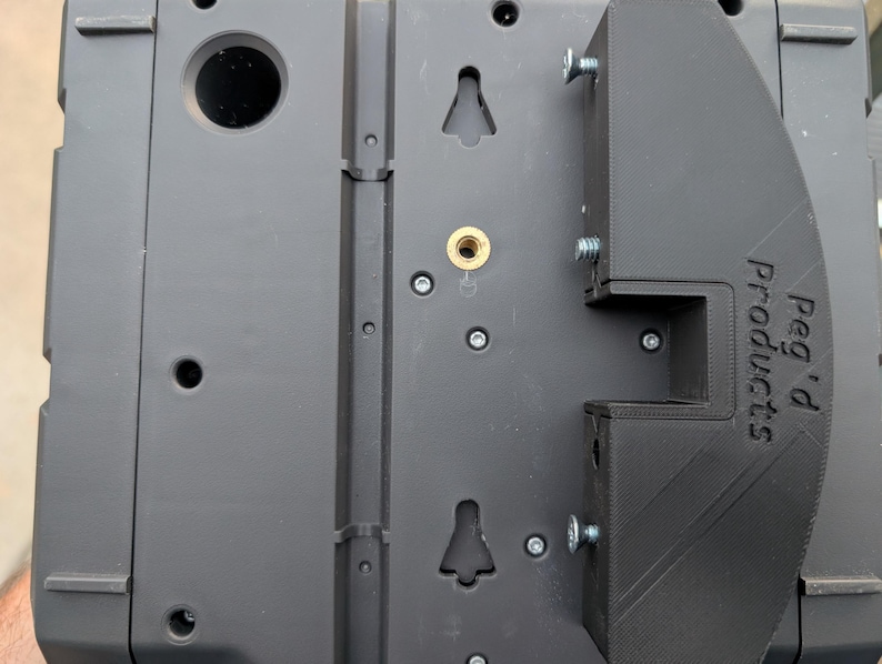May include: Close-up of a black mounting bracket with the text "Peg d Products" printed on the side. The bracket is attached to a gray device with screws. The device has several holes and mounting points.