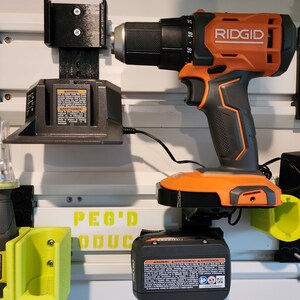May include: A black and orange Ridgid drill with a black battery charger and a black and green Peg'd Products wall mount. The drill has a black and orange handle and a black trigger. The battery charger has a black cord. The wall mount has a black base and a green hook. The drill is mounted on the wall mount. The battery charger is plugged into an outlet. The text "PEG'D PRODUCTS" is visible on the wall mount.