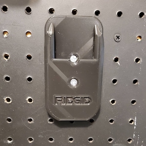 Puede incluir: Soporte de pared de pl&aacute;stico negro con dos puntas y la palabra "RIDGID" impresa en &eacute;l. El soporte est&aacute; fijado a un tablero de clavijas.