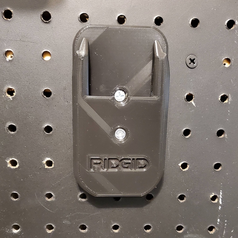 Puede incluir: Soporte de pared de pl&aacute;stico negro con dos puntas y la palabra "RIDGID" impresa en &eacute;l. El soporte est&aacute; fijado a un tablero de clavijas.