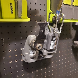 May include: A black and gray oscillating multi-tool with a black blade attached. The tool is hanging on a pegboard with a silver hook. The tool has a label that reads "DCK2012 20V MAX* LITHIUM-ION OSCILLATING TOOL A18L".