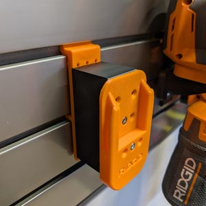 May include: An orange and black power tool holder is mounted on a silver wall. The holder is made of orange plastic and has the brand name "RIDGID" printed on it. The holder is designed to hold a power tool.