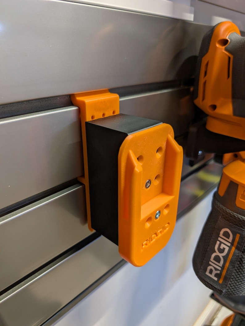 May include: An orange and black power tool holder is mounted on a silver wall. The holder is made of orange plastic and has the brand name "RIDGID" printed on it. The holder is designed to hold a power tool.