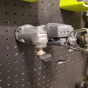 May include: Two black and gray power tools with a silver hook hanging on a pegboard. The tool on the left has a blade attached to the bottom. The tool on the right has the text "RIDGID" printed on the side.