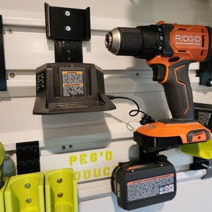 Puede incluir: Un taladro inal&aacute;mbrico negro y naranja con un cargador de bater&iacute;a y una bater&iacute;a de repuesto. El taladro est&aacute; montado en una pared con un soporte negro. El cargador de bater&iacute;a est&aacute; enchufado y montado en la pared con un soporte negro. La bater&iacute;a de repuesto est&aacute; montada en la pared con un soporte negro. La pared es blanca con un tablero de clavijas negro.