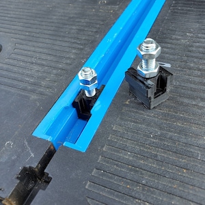 Puede incluir: Dos abrazaderas de metal negro con tuercas y tornillos plateados están unidas a una vía azul. Las abrazaderas se utilizan para asegurar objetos a la vía.