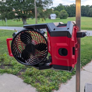 May include: A red and black portable fan with a black fan blade. The fan is mounted on a metal pole. The fan has a red handle and the brand name "Milwaukee" is visible on the side.