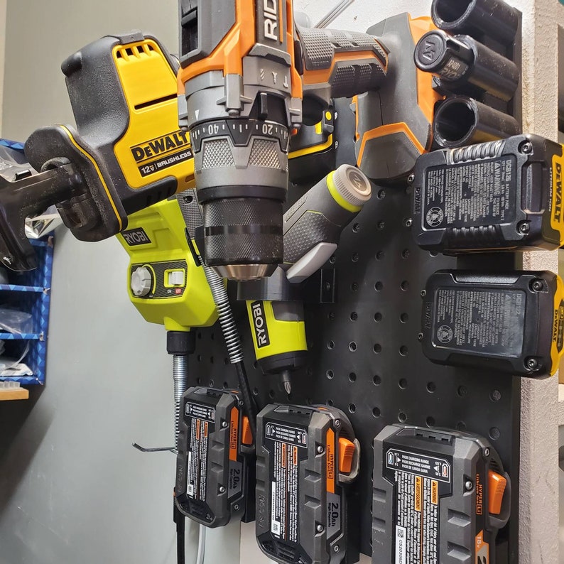 May include: A pegboard with various power tools and batteries hanging on it. The tools include a yellow and black DeWalt drill, a black and green Ryobi drill, a black and orange Ryobi drill, and a black and yellow DeWalt battery. The batteries are labeled "2.0Ah" and "DCB125".