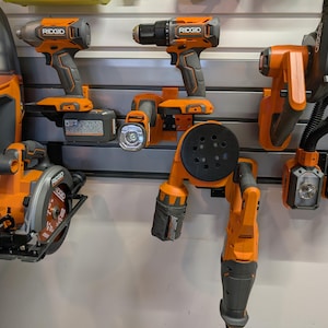May include: An assortment of orange and gray power tools, including a circular saw, drill, and sander, are mounted on a wall. The tools are from the brand Ridgid. The tools are displayed on a gray wall.