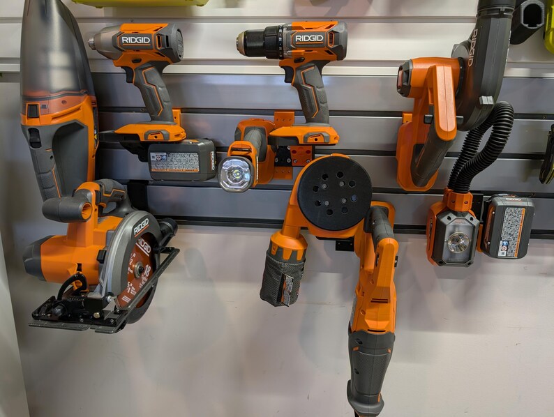 May include: An assortment of orange and gray power tools, including a circular saw, drill, and sander, are mounted on a wall. The tools are from the brand Ridgid. The tools are displayed on a gray wall.