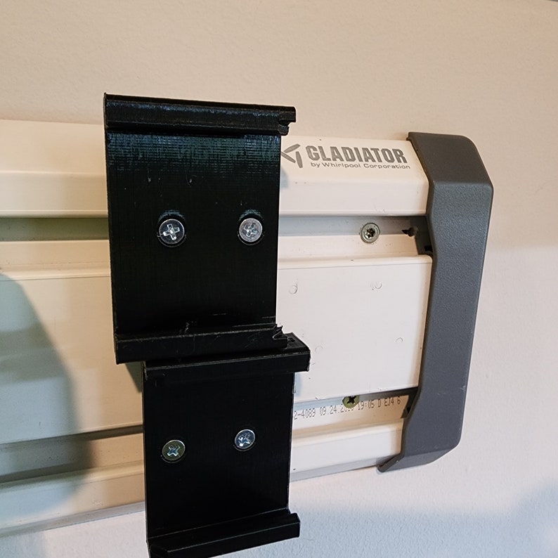 May include: Two black plastic wall mount brackets with screw holes. The brackets are attached to a white Gladiator wall storage system with the text "GLADIATOR by Whirlpool Corporation".
