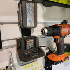May include: A black and orange cordless drill with a black battery charger and a black and orange battery pack. The drill is mounted on a wall-mounted pegboard with the text "PEG'D PRODUCTS" printed on it. The drill has the text "RIDGID" printed on it.