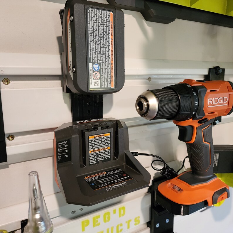 May include: A black and orange cordless drill with a black battery charger and a black and orange battery pack. The drill is mounted on a wall-mounted pegboard with the text "PEG'D PRODUCTS" printed on it. The drill has the text "RIDGID" printed on it.