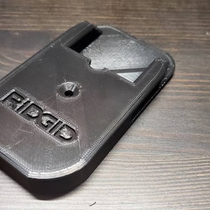 Puede incluir: Soporte de pared de pl&aacute;stico negro impreso en 3D con la palabra "RIDGID" en relieve en la parte delantera. El soporte tiene una ranura para una herramienta y un agujero para un tornillo.