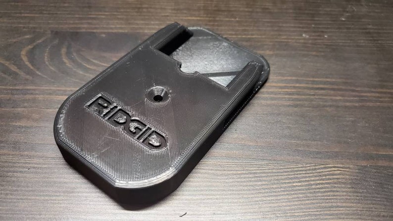 Puede incluir: Soporte de pared de pl&aacute;stico negro impreso en 3D con la palabra "RIDGID" en relieve en la parte delantera. El soporte tiene una ranura para una herramienta y un agujero para un tornillo.