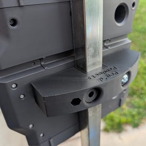 May include: Close-up of a black plastic product with the text "Peg d. Products" printed on it. The product is attached to a metal pole with screws. The background is blurred, showing green grass and an orange object.