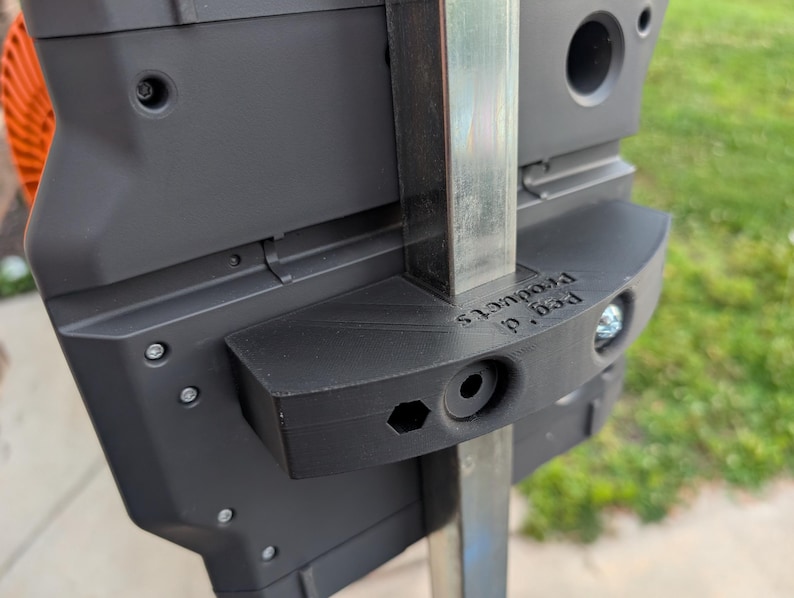 May include: Close-up of a black plastic product with the text "Peg d. Products" printed on it. The product is attached to a metal pole with screws. The background is blurred, showing green grass and an orange object.