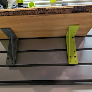 Puede incluir: Una estantería de madera sostenida por dos soportes metálicos. Un soporte está pintado de verde lima, el otro es gris oscuro. La estantería está montada en una pared con ranuras horizontales. La estantería es de madera.