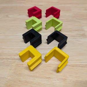 May include: Eight L-shaped plastic corner protectors are shown, arranged in pairs. Each pair is a different colour: red, green, black, and yellow. The objects are on a light wood-grain surface. The protectors are designed to protect corners.
