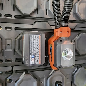 Puede incluir: Una luz de trabajo inal&aacute;mbrica negra y naranja con una manguera flexible y una bater&iacute;a con las etiquetas "RIDGID 18V", "WARNING", "AVERTISSEMENT", "ADVERTENCIA" y "RICOH".
