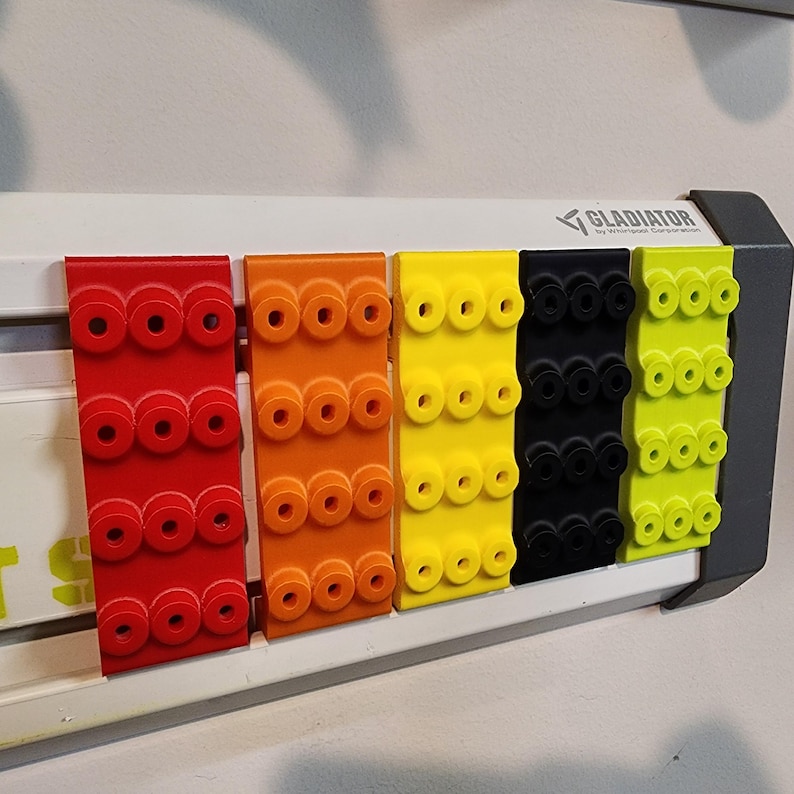 May include: A row of colorful Gladiator brand storage clips in red, orange, yellow, black, and lime green. Each clip has multiple circular holes for hanging items. The clips are mounted on a white and gray metal frame.