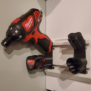 Puede incluir: Un taladro inalámbrico Milwaukee rojo y negro con una batería conectada está montado en un sistema de almacenamiento de pared Gladiator. El taladro está colgado de un soporte de plástico negro con un soporte de batería de plástico negro.